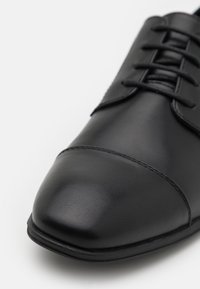 Chaussure habillée en cuir noir au fini lisse, à bout pointu et à lacets. Présente une couture discrète et un design élégant et raffiné.