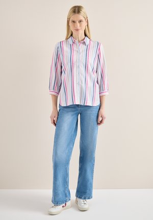 Nainen seisoo, hänellä on päällään pastelliraidallinen nappisuljettava paita, jossa on kolmen neljänneksen hihat, vaaleansiniset leveälahkeiset farkut ja valkoiset tennarit.