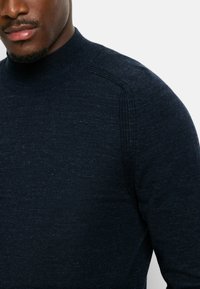 Dunkelblauer Strickpullover mit hohem Ausschnitt, gerippter Textur und subtilen Farbklecksen in helleren Tönen. Besitzt eine Schulterdetails.