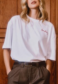 Kobieta ubrana w białą, oversize'ową koszulkę z czerwonym napisem "House of Sunny", brązowe spodnie z wysokim stanem oraz czarny pasek, stojąca oparta o drewnianą ścianę.