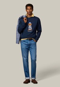 Mand iført marineblå sweatshirt med hundegrafik, gul skjorte med krave, blå jeans, brune sko, der holder en blåternet jakke over skulderen.