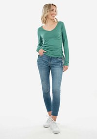 Camicia verde a maniche lunghe con scollo a V, abbinata a jeans skinny blu con strappi. Il modello indossa sneakers platform bianche.