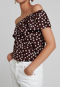 Ruskea pilkullinen olkaimeton toppi, jossa on röyhelökaulus, valmistettu kevyestä kankaasta, yhdistettynä valkoisiin korkeavyötäröisiin housuihin.