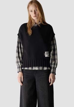 Young woman wearing a black knit vest over a gray plaid long-sleeve shirt and loose black pants standing against a gray background.