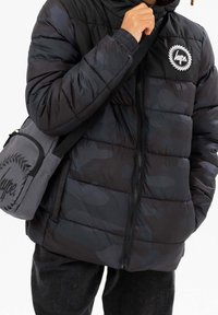 Czarna kurtka puchowa z kapturem, z pikowanym wzorem, elastycznymi mankietami i białą naszywką z logo. Towarzyszy jej szara torba na ramię.