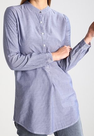 Femme portant une chemise à manches longues, bleu clair et blanc à fines rayures, avec col mandarin, partiellement rentrée dans un jean en denim.
