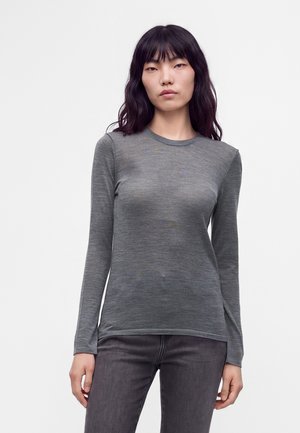 Femme aux longs cheveux foncés portant un haut gris ajusté à manches longues et un pantalon gris foncé sur un fond clair uni.