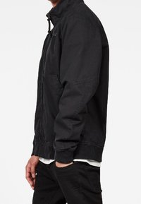 Man i svart zip-up jacka över en vit skjorta med svarta byxor, stående med en hand i fickan.