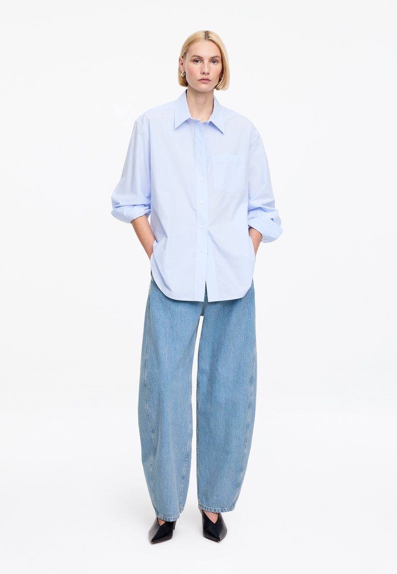 Mujer rubia de pie con las manos en los bolsillos, vistiendo una camisa azul claro de botones extragrande, vaqueros de pierna ancha y zapatos negros puntiagudos.