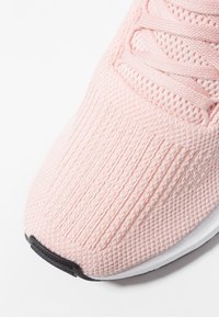 Vaaleanpunainen urheilukenkä, jossa on teksturoitu verkkopäällinen, pystysuora uurrekuvio ja tasaiset nauhat. Kenkässä on valkoinen pohja, jossa musta korostus kärjessä.