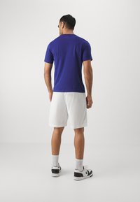 Camiseta morada de manga corta combinada con pantalones cortos de malla blanca y zapatillas en blanco y negro. El conjunto es casual y deportivo.