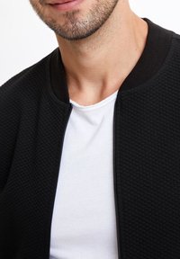 Czarna kurtka bomberka z pikowaną fakturą, zapinana na zamek, z prążkowanym kołnierzem, noszona na białej koszulce.