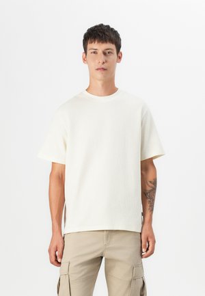 Korte mouwen, lichtbeige katoenen t-shirt met een ribbelstructuur, ronde hals en zijslits, gedragen met beige cargobroeken.