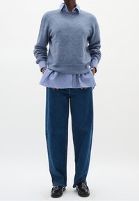 Personne portant un pull bleu col en V sur une chemise rayée bleue et blanche, un jean large bleu foncé et des mocassins noirs, debout les mains dans les poches.