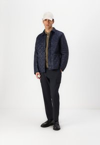 Jachetă matlasată bleumarin cu un design cu guler, purtată cu o cămașă măslinie, pantaloni negri și pantofi negri. O șapcă de culoare deschisă completează ținuta.
