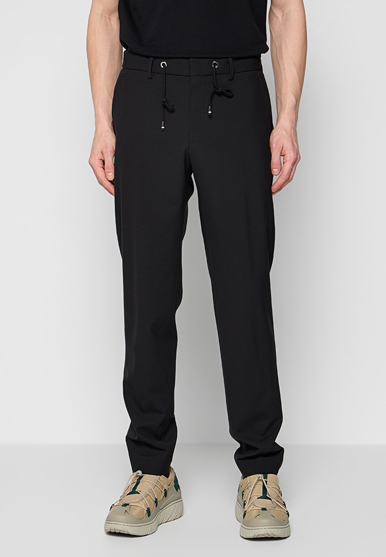 Pantalons noirs fuselés avec taille élastique à cordon de serrage et poches latérales, associés à des chaussures beiges avec des accents verts et un dessus texturé.