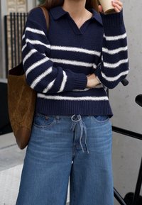 Námořnicky modrý pletený svetr s bílými pruhy, výstřihem do V a žebrovanými manžetami; kombinován s širokými džíny světle modré barvy a hnědou taškou.