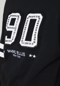 Maglietta sportiva nera con un grande "90" in bianco e argento con borchie sul davanti, con ricamato "MARC ELLIS NEW YORK" sotto e strisce bianche sulle maniche.