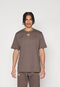 T-shirt en coton marron à manches courtes, avec un logo jaune sur la poitrine. Le tissu semble lisse et décontracté dans son design.