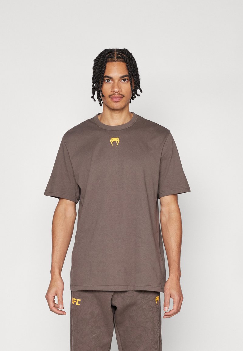 T-shirt en coton marron à manches courtes, avec un logo jaune sur la poitrine. Le tissu semble lisse et décontracté dans son design.
