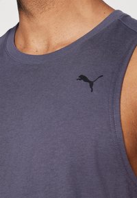 Chemise sans manches violette en coton, avec un col rond et un petit logo noir de Puma sur la poitrine gauche. Texture lisse.