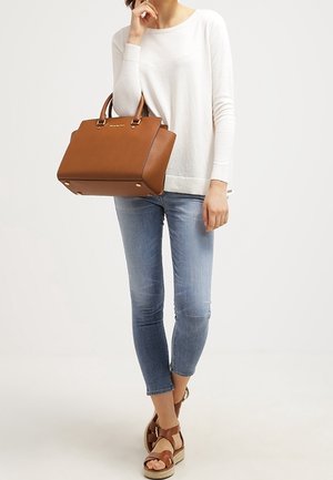 Hnědá kožená kabelka se strukturovaným designem, zlatými kovovými doplňky a dvěma horními rukojetmi. Modelka má na sobě bílý top a modré úzké džíny.