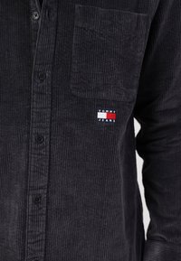 Camicia in corduroy grigio scuro con design a bottoni, taschino sul petto e un piccolo cartellino con il marchio in accenti rossi, bianchi e blu.