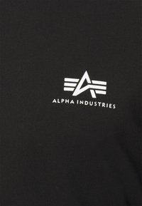 Schwarzes T-Shirt aus Baumwolle mit einem weißen Alpha Industries-Logo, das stilisierte Buchstaben zeigt, und drei horizontalen Streifen über dem Logo.