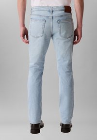 Gaiši zilas džinsa bikses ar šauru piegriezumu, piecu kabatu dizainu un brūnu ādas aizmugures plāksnīti. Tām ir taisnas kājas un smalka tekstūra.