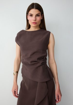 Femme aux longs cheveux bruns portant un top marron foncé sans manches et une jupe, debout devant un fond uni clair.