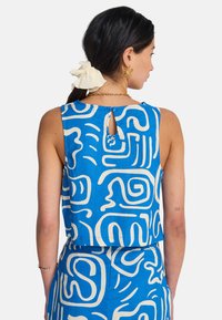 Μπλε και λευκό τοπ με μοτίβα και cropped σχεδιασμό, με αφηρημένες σπείρες και λεπτομέρεια πίσω με άνοιγμα σε σχήμα κλειδαρότρυπας. Κατασκευασμένο από ελαφρύ ύφασμα.