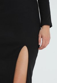 Černé žebrované šaty s dlouhým rukávem, které mají vysoký rozparek po boku. Měkká textura, fitted design a minimalistický styl.