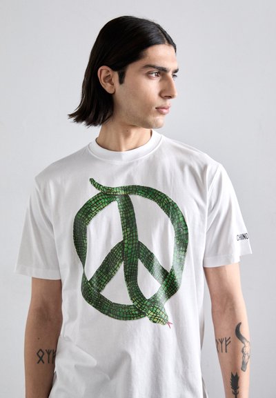 Camiseta de algodón blanca con un gran diseño de serpiente verde formando un símbolo de paz; mangas cortas, cuello redondo y detalle de la marca en la manga.
