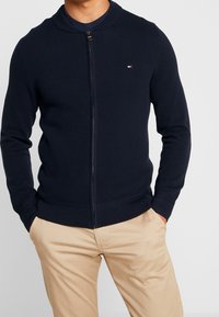 Navy modrý pletený svetr na zip s texturovaným vzorem, ribkovaným límcem a manžetami, s malým logem na prsou, doplněný béžovými kalhotami.