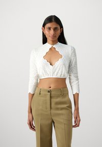 Biely čipkovaný cropped top s vlnkovaným výstrihom a dlhými rukávmi, doplnený vysokými béžovými nohavicami.