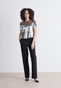 Zilveren pailletten top met off-the-shoulder ontwerp, gecombineerd met zwarte pantalons met streepjes. Model draagt zwarte puntige hakken. Effen achtergrond.