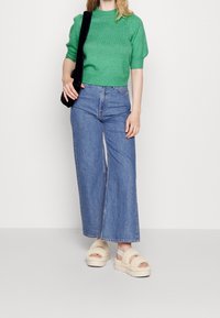 Groene gebreide trui met korte mouwen, lichte blauwe wijde jeans en beige donzige sandalen met een gestructureerde band. Zwarte tas over één schouder.