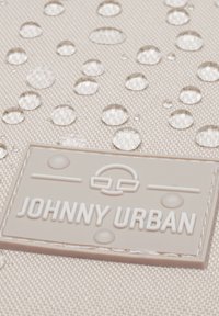 Gouttelettes d’eau sur un tissu texturé beige au-dessus d’un écusson en caoutchouc Johnny Urban, mettant en avant la résistance à l’eau du matériau.