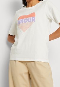 Vit bomullströja med en hjärtformad grafik i korall och lila med ordet "AMOUR." Korta ärmar och en avslappnad passform.