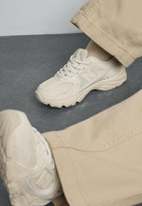 Hvite mesh-sneakers med teksturerte detaljer, polstret såle, rund t�å og snøring, brukt sammen med beige bukser på en grå overflate.