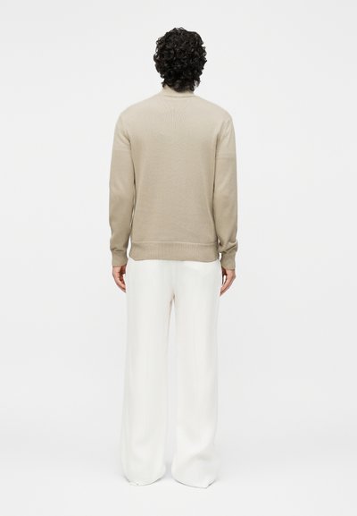 Isik, kellel on lokkis tumedad juuksed, kandes beeži pikkade varrukatega kampsunit ja laiu valgeid pükse, seisab valgel ühevärvilisel taustal seljaga vaataja poole.