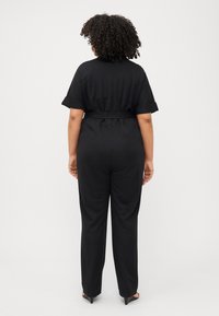 A person with curly hair stands facing away, wearing a fitted black jumpsuit and heels, against a plain white background.