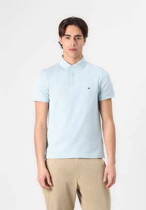 Lys blå polo skjorte, bomullsmateriale, med knappeskjorte, korte ermer, med en liten logo på venstre bryst. Båret sammen med beige bukser.