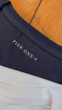 Crna pamučna košulja s rebrastim ovratnikom na kojem se nalazi bijela oznaka "PIER. ONE". Bijela košulja vidljiva ispod. Prirodna drvena pozadina.
