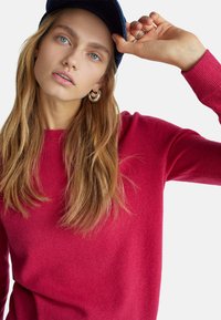 Maglione lavorato a maglia di un vivace rosa, con scollatura tonda, maniche lunghe e una texture morbida, abbinato a un berretto blu scuro e orecchini a cerchio argento.