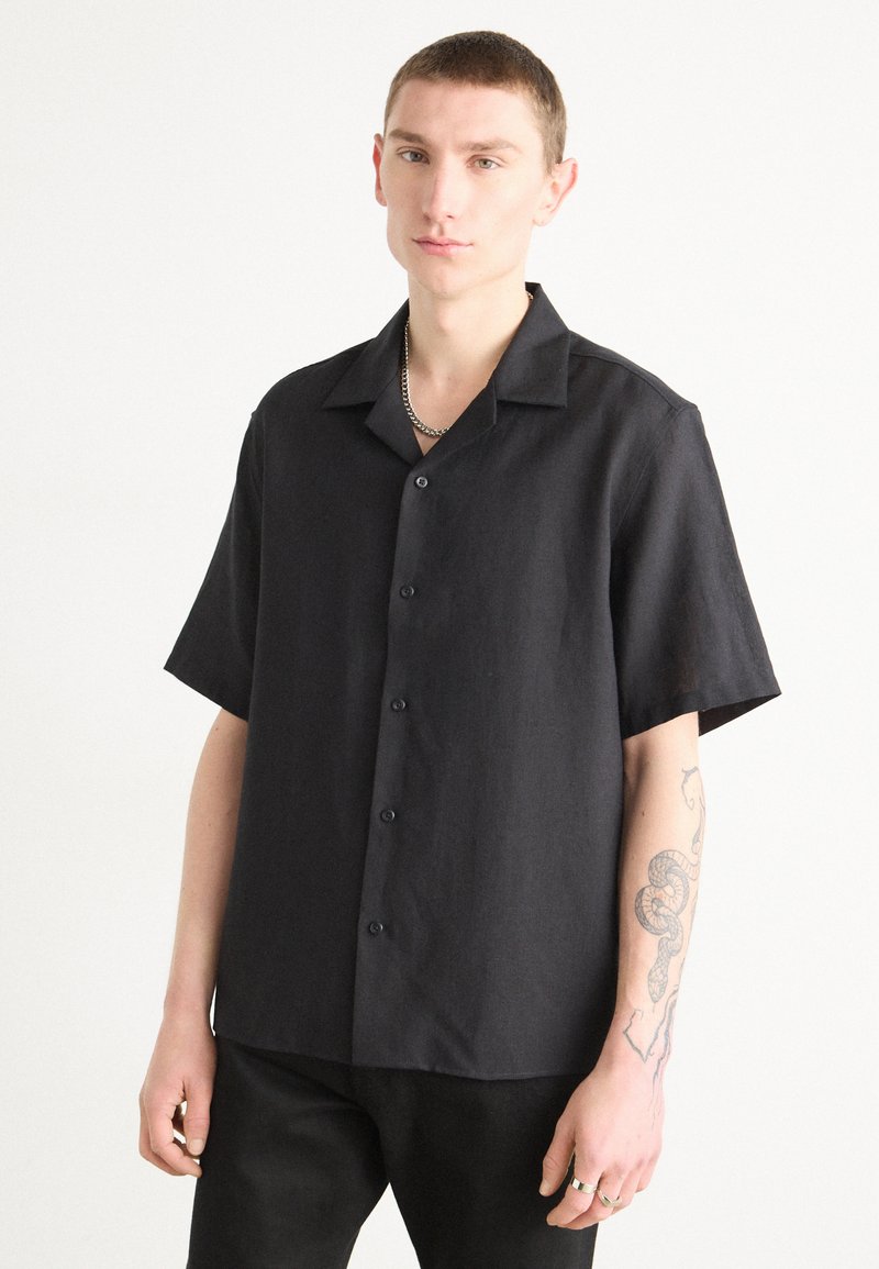 Camisa de botones negra de manga corta hecha de tela texturizada, con cuello y botones negros, combinada con pantalones negros.