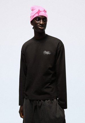 Homme portant un bonnet rose vif et un t-shirt noir à manches longues avec un texte blanc sur la poitrine, debout devant un fond clair uni.