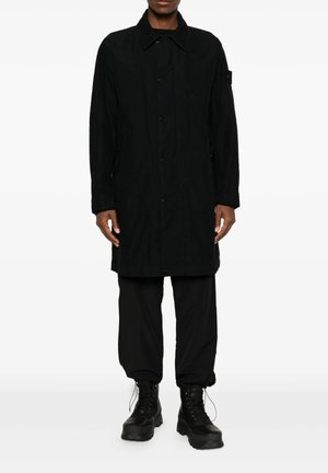 Uomo che indossa un lungo cappotto nero, pantaloni neri e stivali neri con lacci, in piedi davanti a uno sfondo bianco.