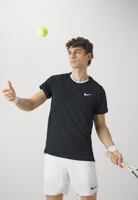 Svart trenings T-skjorte med hvit Nike-logo, kombinert med hvite shorts. Ung mann står, kaster en lys grønn tennisball, holder en racket.
