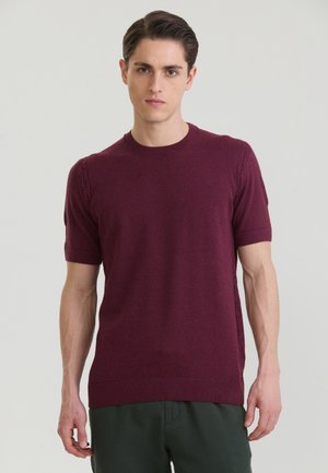 Fiatal férfi, rövid barna hajjal, bordó rövid ujjú kötött pulóvert és sötétzöld nadrágot viselve, egyszínű háttér előtt áll.